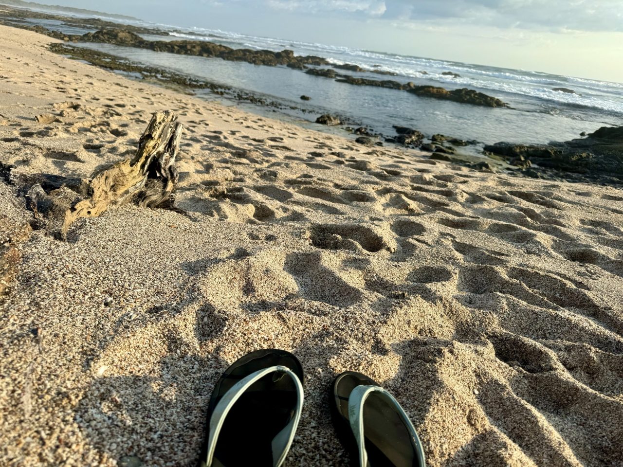 Driftwood on a wide beach at low tide, rocks and surf in the distance, a pair of shoes in the sand