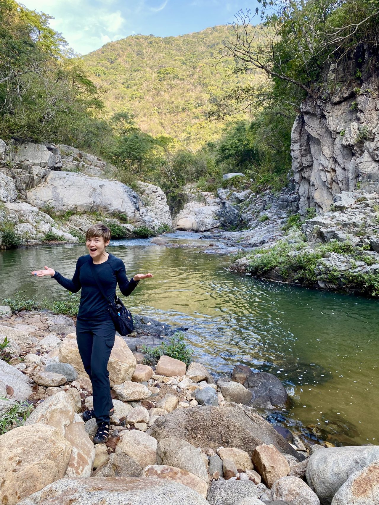 Masha standing on boulders beside a clear green creek below a cliff