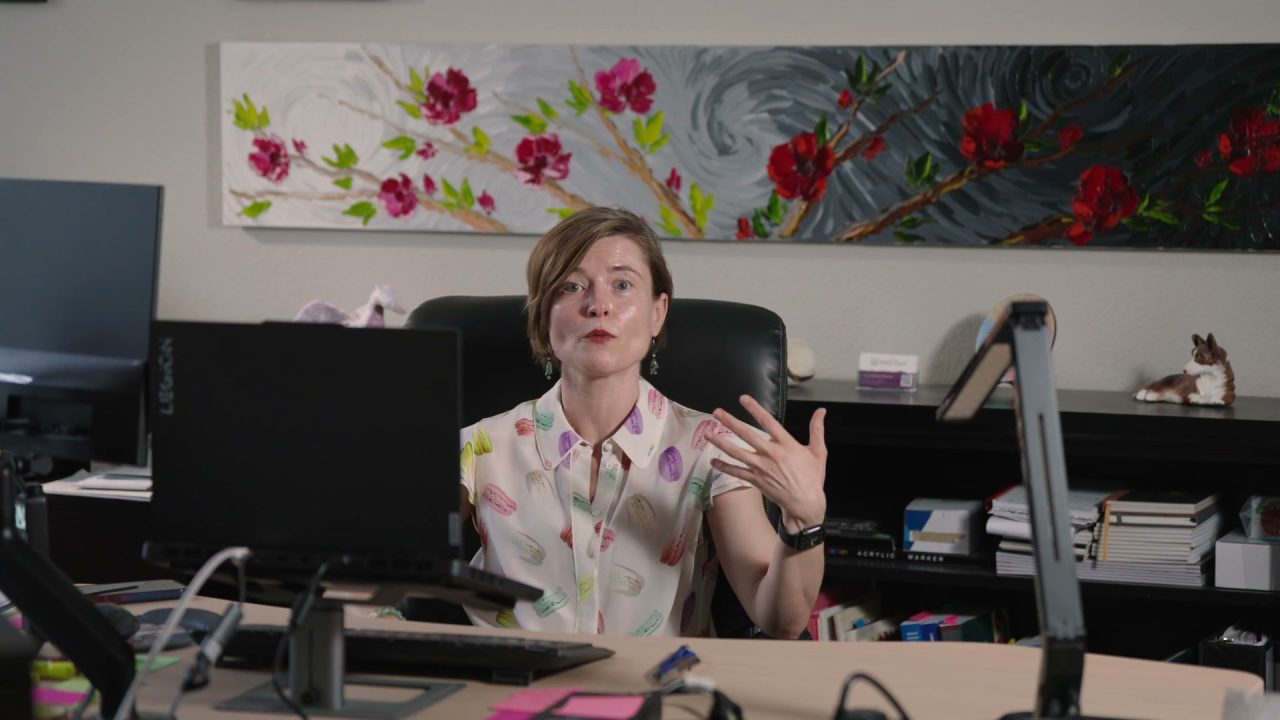 Masha at her desk, mid-sentence, one hand raised as she explains something