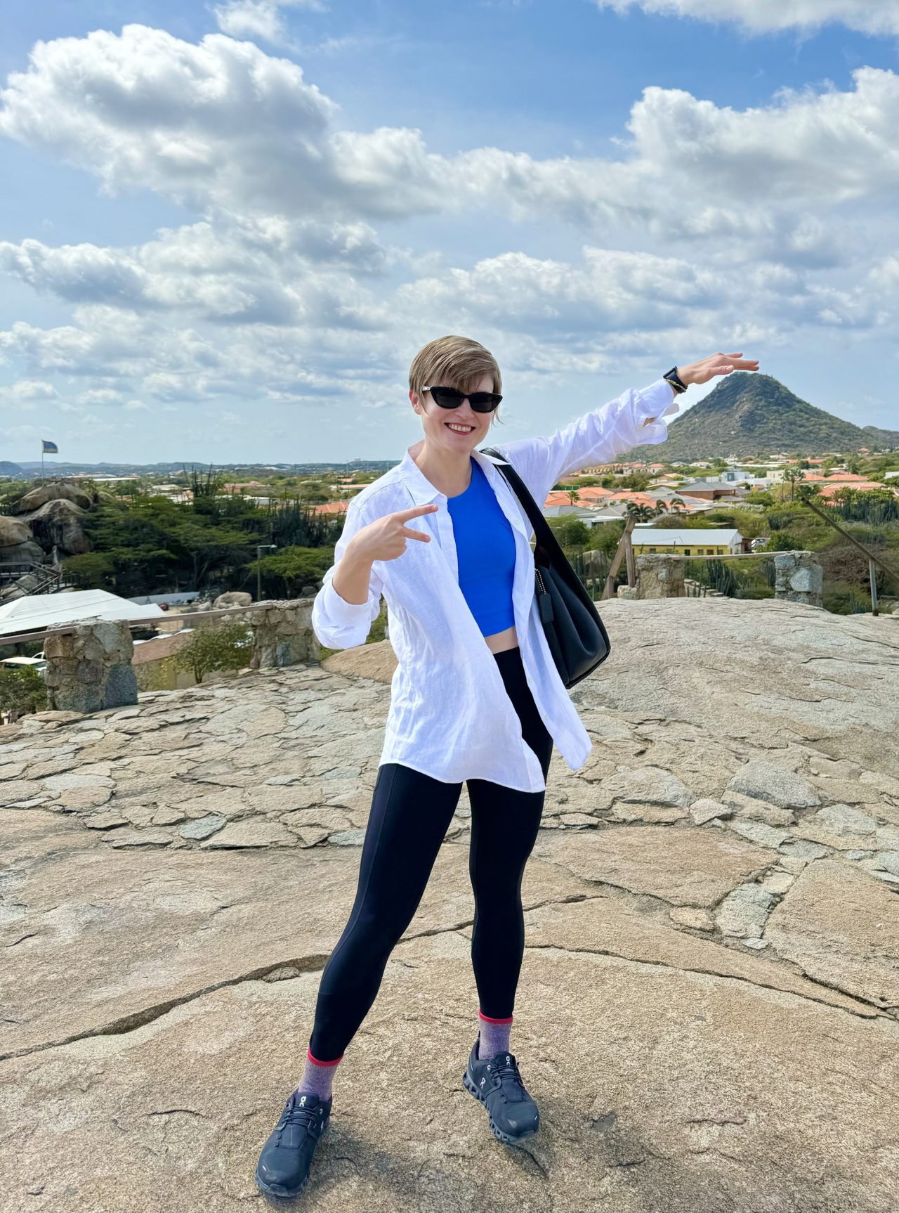 Masha on a rocky hilltop in a white jacket, pointing at the view behind her