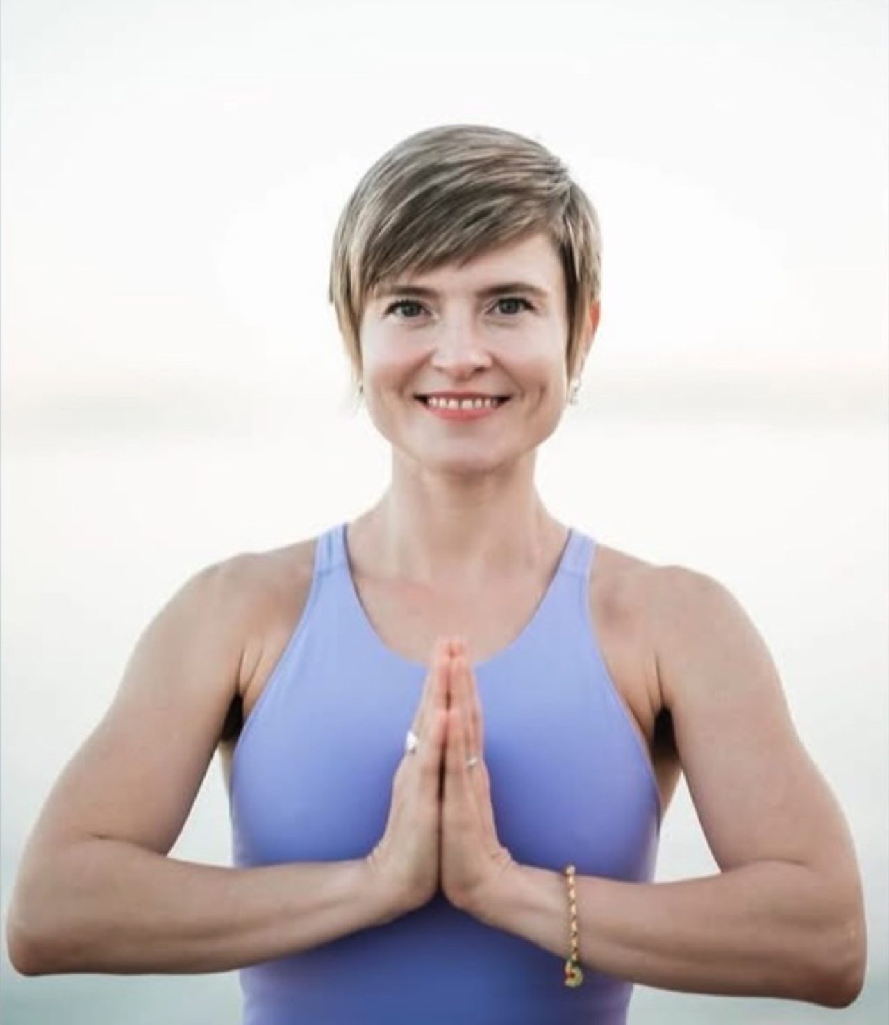 Masha in a lavender top with her palms together at her chest, the sea behind her