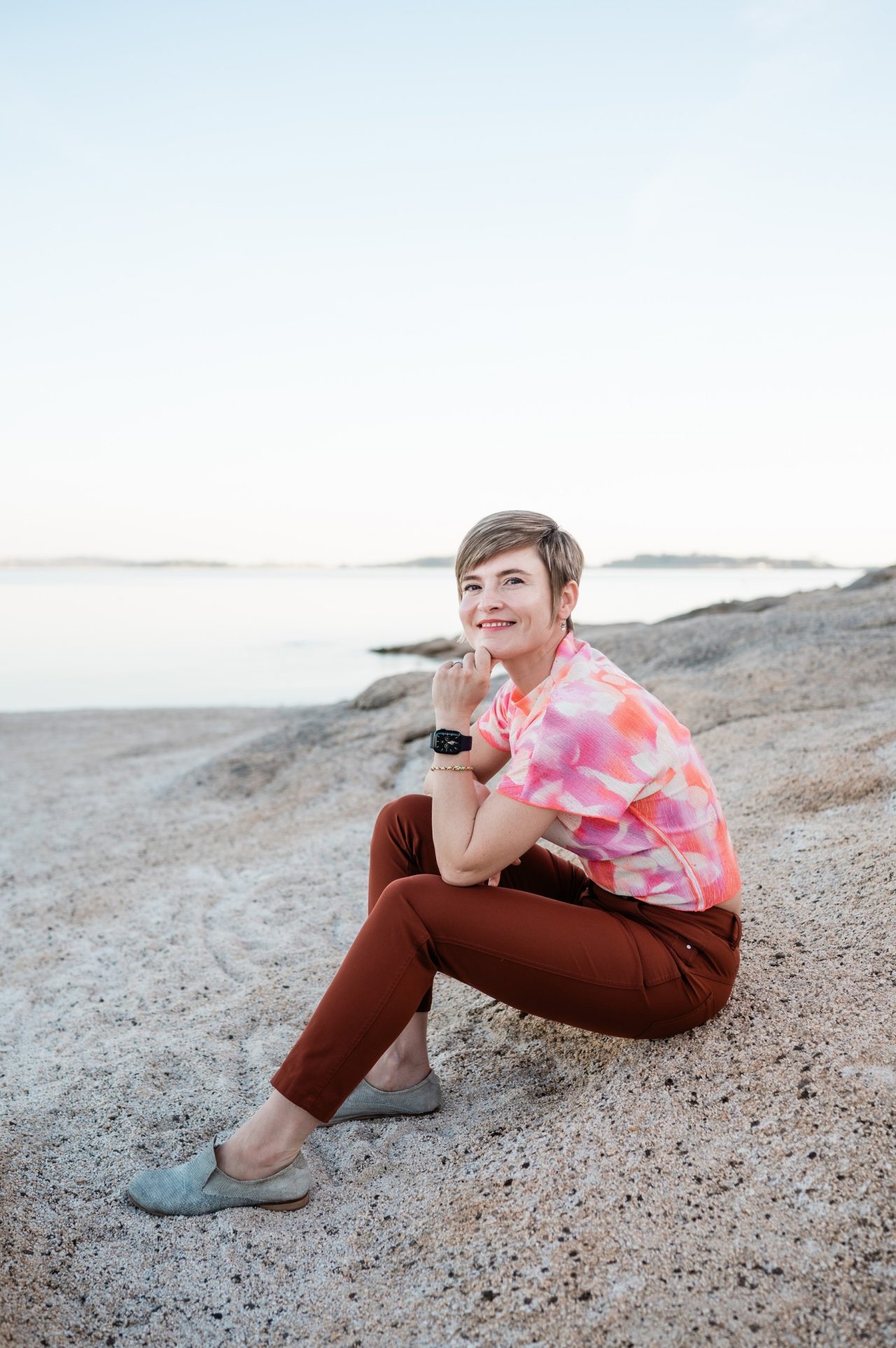 Masha seated on a pale granite shore, chin on her hand, in a tie-dye top