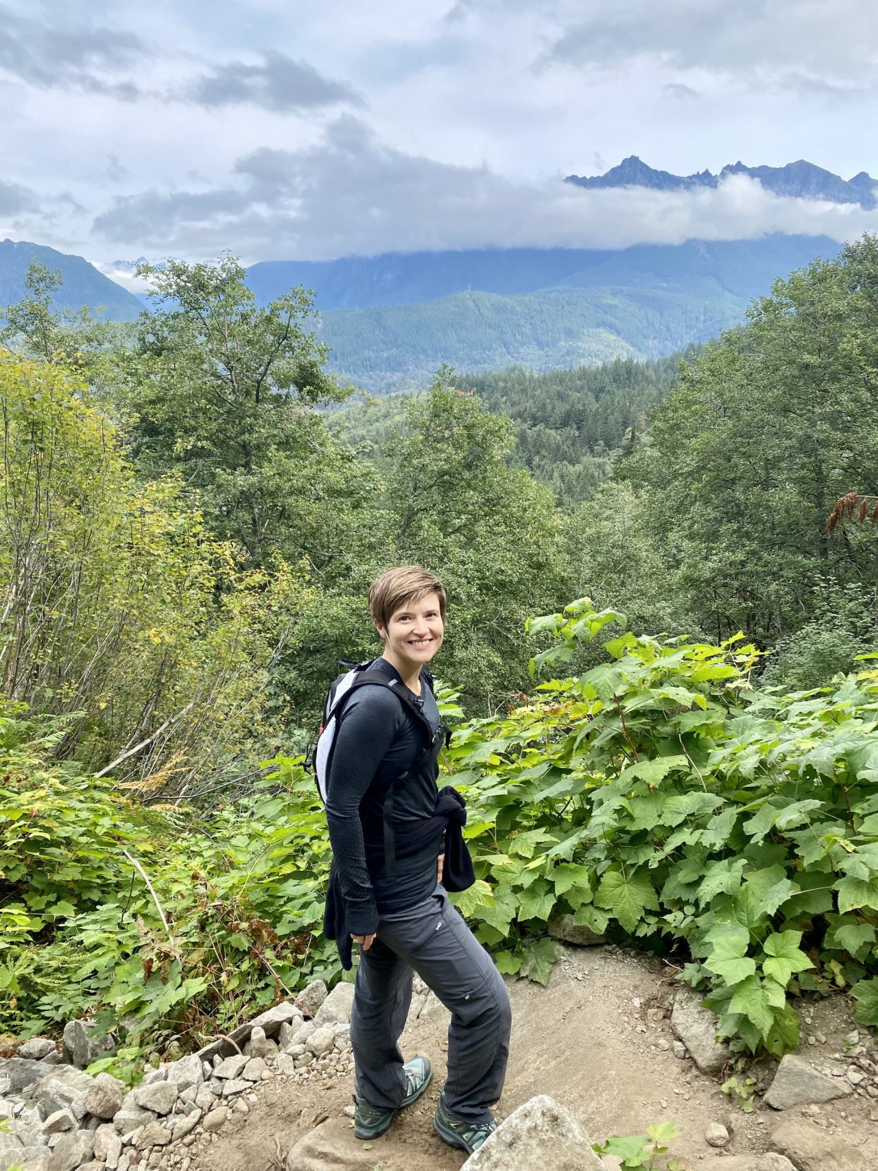 Masha on a mountain trail among ferns, with a valley and peaks behind her