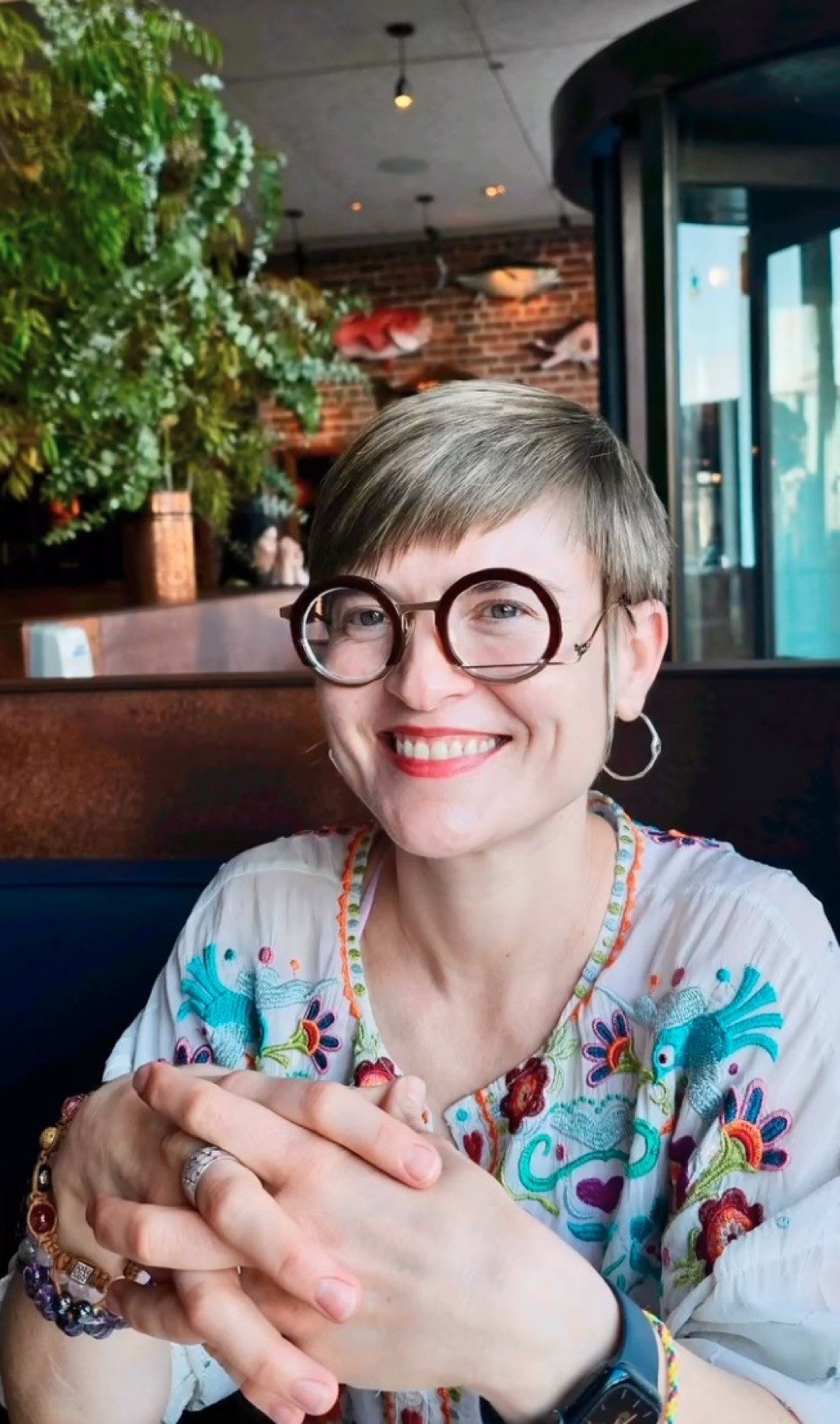 Masha in round glasses and a floral top, hands clasped, smiling
