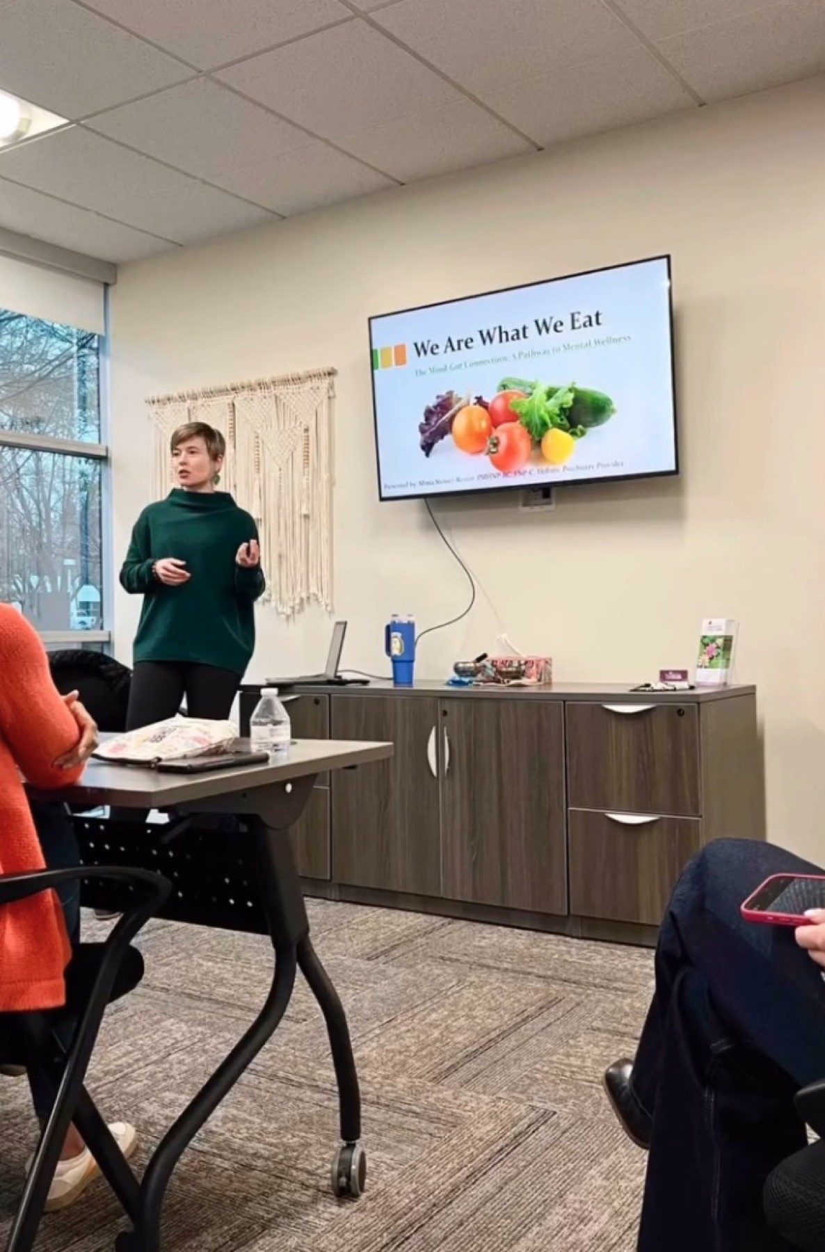 Masha presenting to a seated group in a meeting room, a slide on the screen behind her
