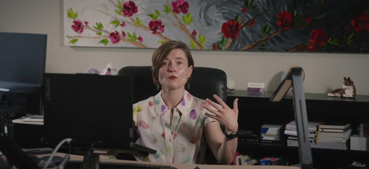 Masha talking at her desk in her Lakewood office, a floral painting on the wall behind her