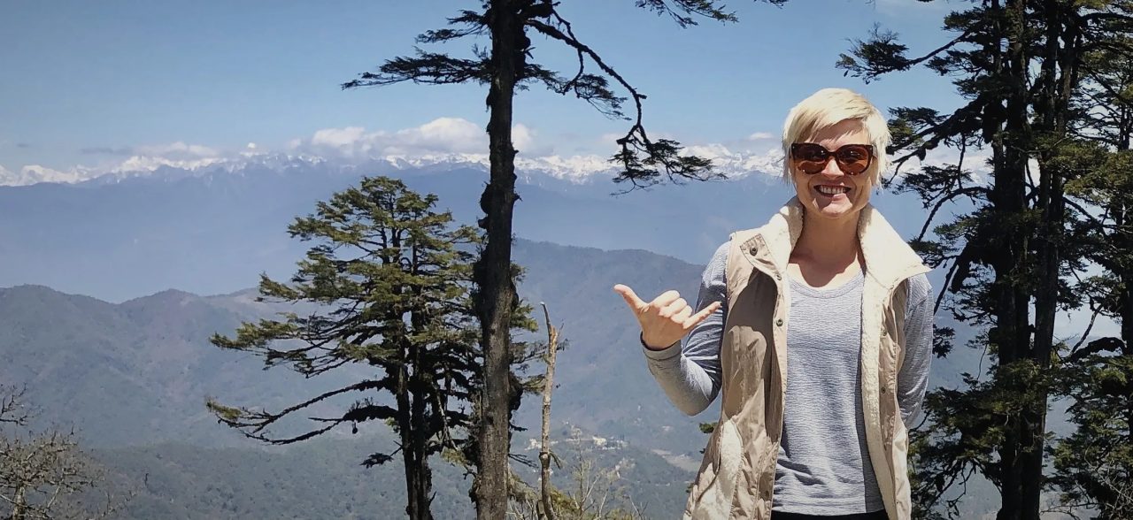 Masha smiling in sunglasses and a vest on a mountain overlook, snowy peaks behind her