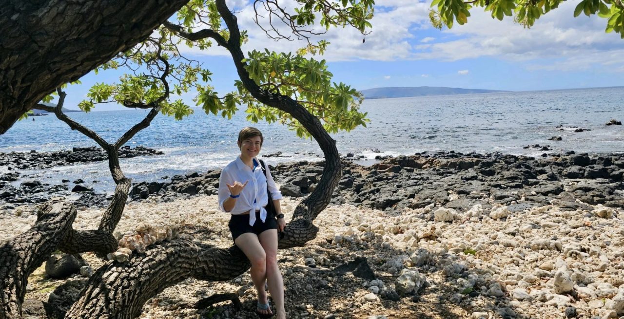 Masha Steiner-Renoir sitting on a low tree branch above a rocky shoreline, the ocean behind her.
