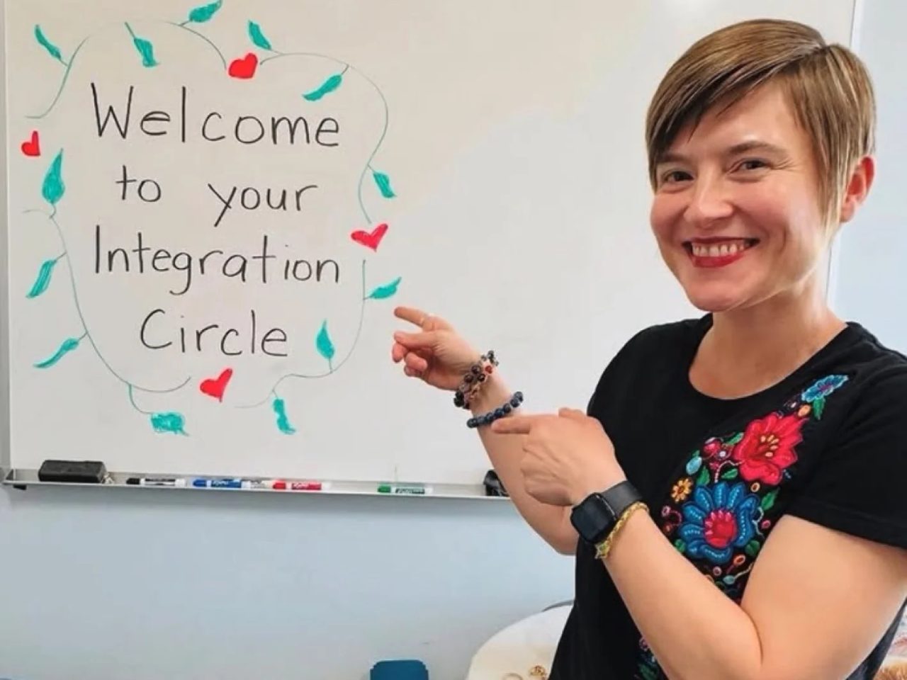 Masha Steiner-Renoir pointing to a whiteboard that reads Welcome to your Integration Circle, floor cushions stacked beside her.