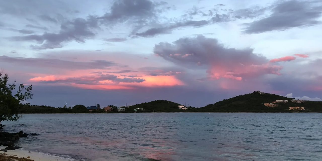 Pink and grey storm clouds over a quiet bay with low green hills on the far shore