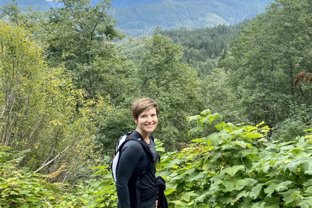 Masha Steiner-Renoir on a mountain trail with a daypack, looking back over her shoulder, forested ridges under cloud behind her.