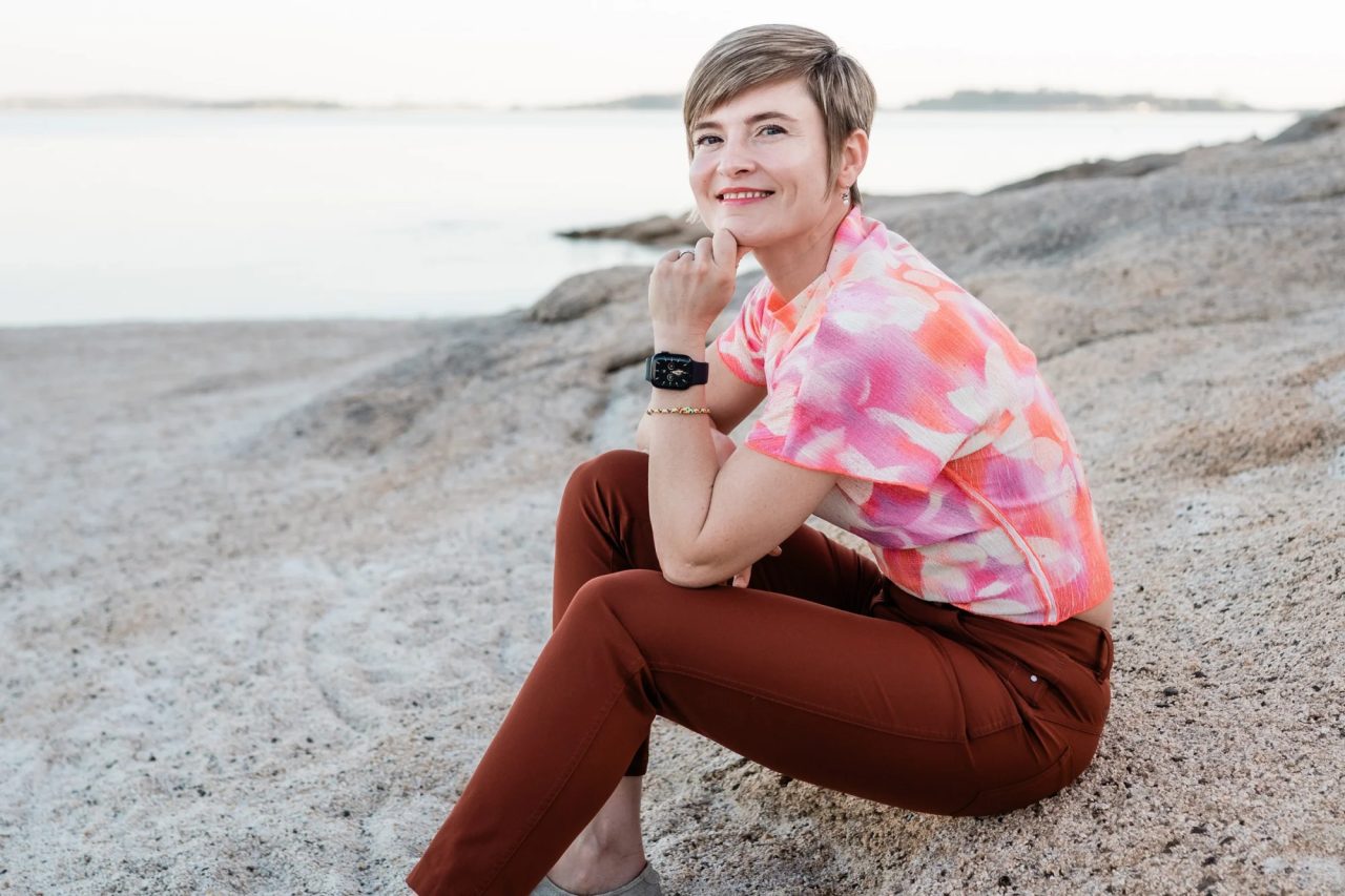Masha Steiner-Renoir sitting on a granite shore at dusk with her chin resting on her hand, still water behind her.