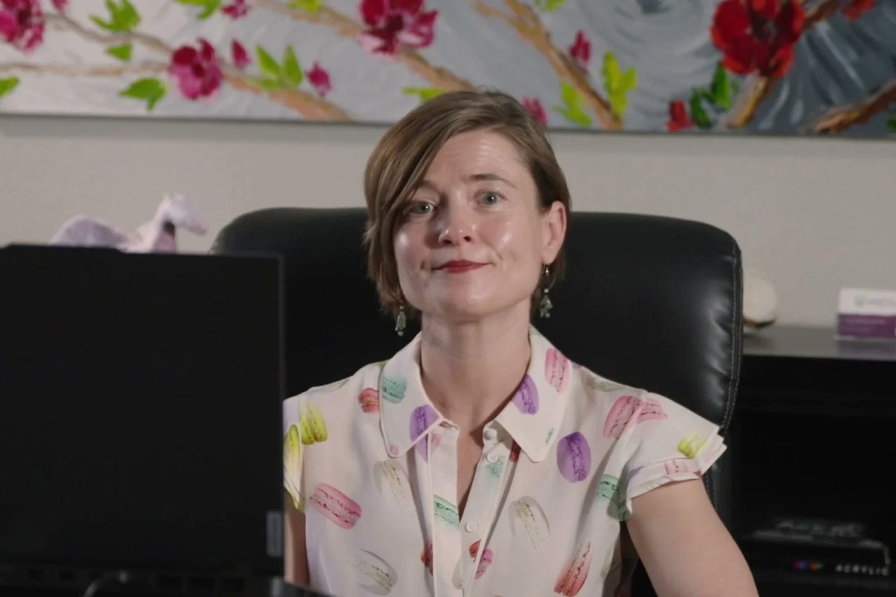 Masha Steiner-Renoir seated at the desk in her office, a painting of red blossoms on the wall behind her.